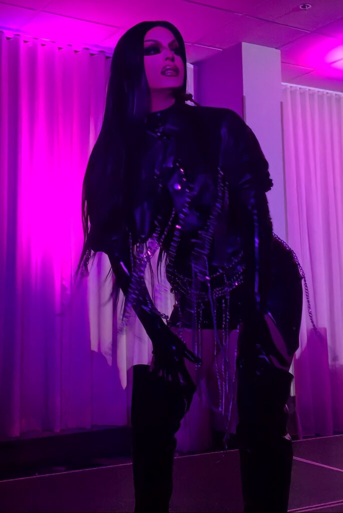 Alt text: A Drag queen with long black hair is wearing a black heeled boots in a leather jacket and shorts that are fringed with metal chains.