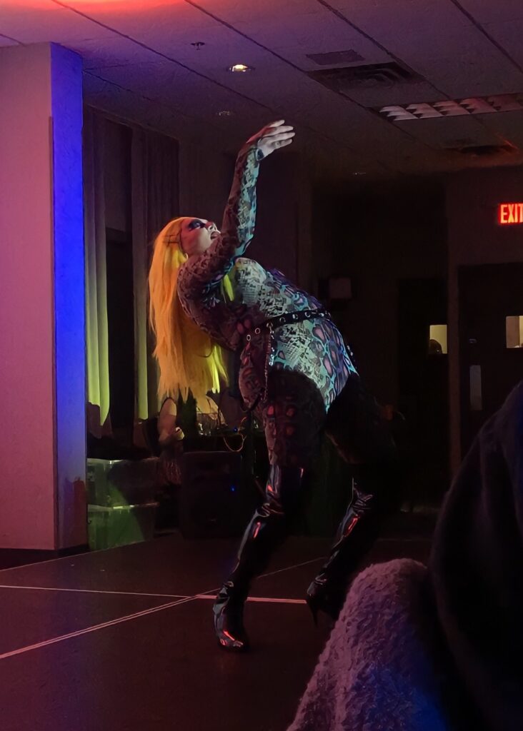 Alt text: A Drag queen with long straight yellow hair is wearing a green and purple leopard print body suit, with a black leather studded belt and latex heeled boots. Her back is arched as she goes into a backbend.