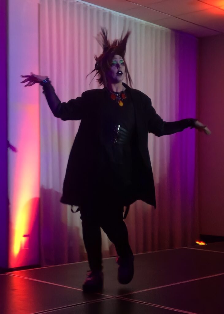 Alt text: A Drag prince with spiky red hair is prancing across the stage in a long, black overcoat. 