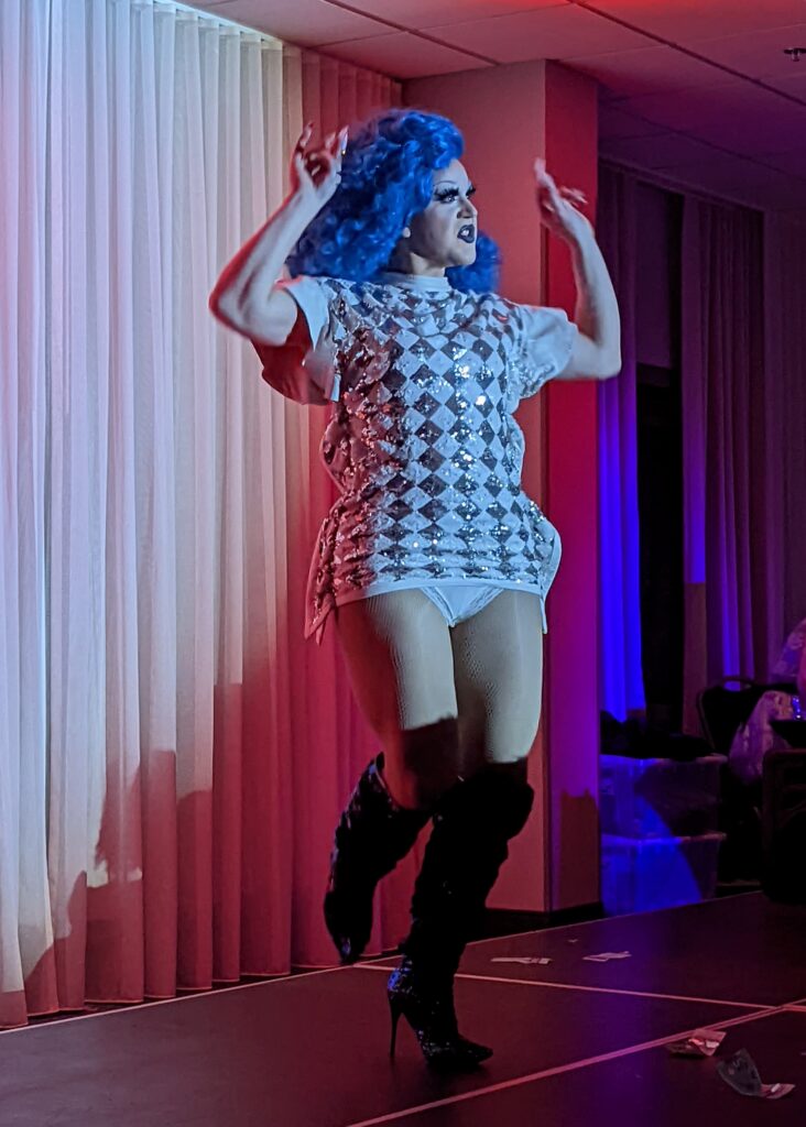 Alt text: A Drag queen with curly, cobalt hair is wearing a white t-shirt dress with a silver diamond pattern across and black latex heeled boots. She is dancing with her arms raised and her right leg popped up