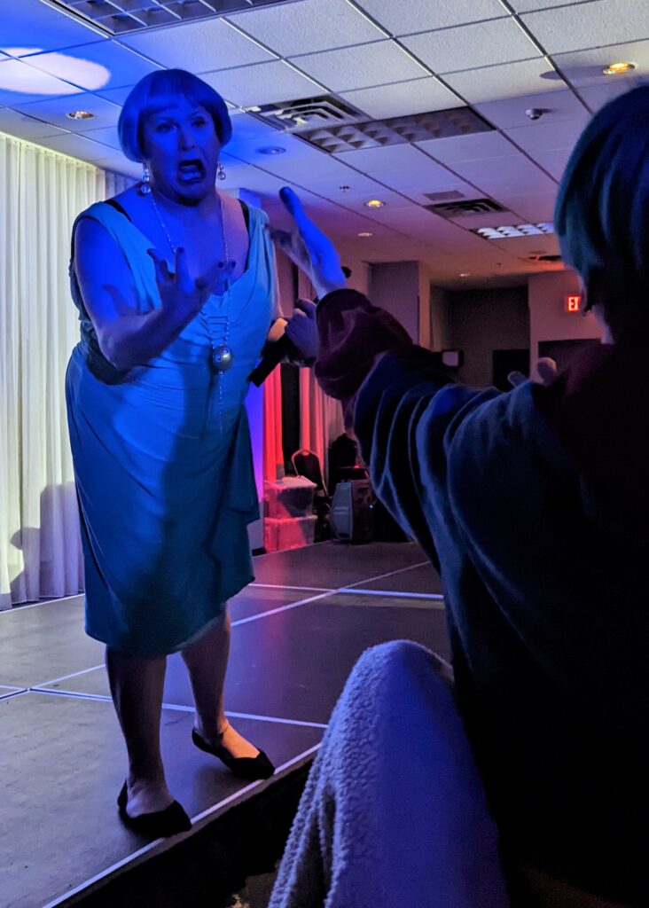 Alt text: A Drag queen with a short bob haircut wears a blue dress and golden bobble earrings with a necklace to match. She is cast in blue lighting as she serenades an audience member.
