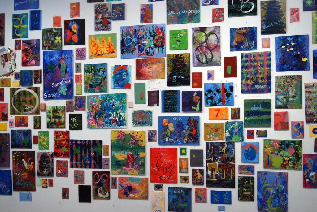 A white wall covered by brightly coloured paintings of various sizes.