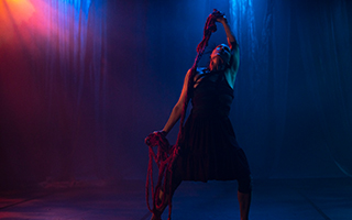 A performer in Firewater Thunderbird Rising 2 - Friday Creeations. Blue and pink lights are in the background and the performer is posed with a rope.