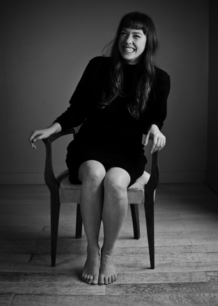 A black and white photo of Tracey Guptill smiling while sitting on a chair in a dress against a plain background. 