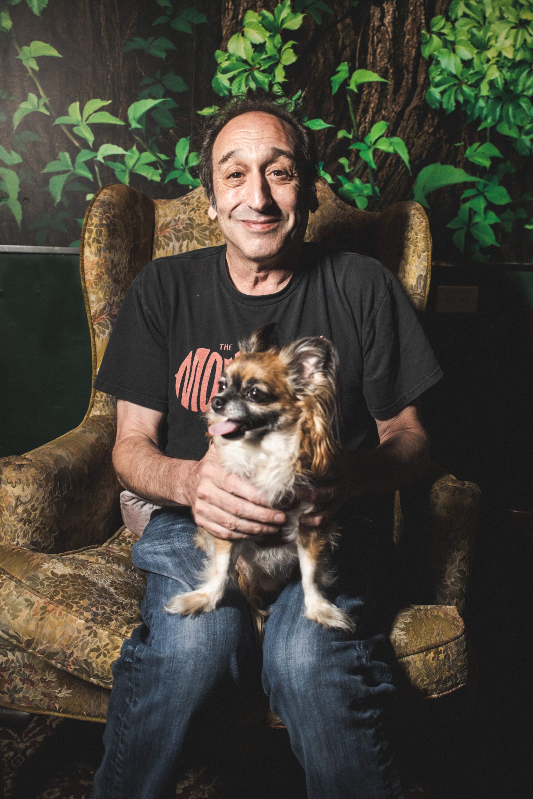 Jeremy Hotz sits on a chair with a dog on his lap in front of a tree.