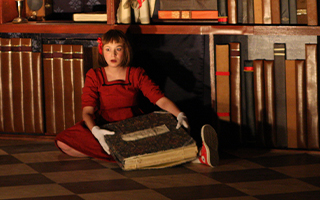 A young girl sits in front of a large bookshelf holding a large book. She wears a red dress and white gloves. Text in the bottom right corner reads: "Grand OnStage"