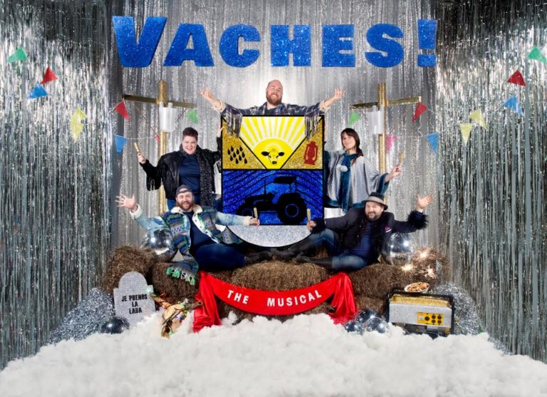 A background of silver streamers and fake snow on the ground. Atop the snow are hay bales with five people sitting on them. One cross is on each side of the group. Text reads: "Vaches The Musical"