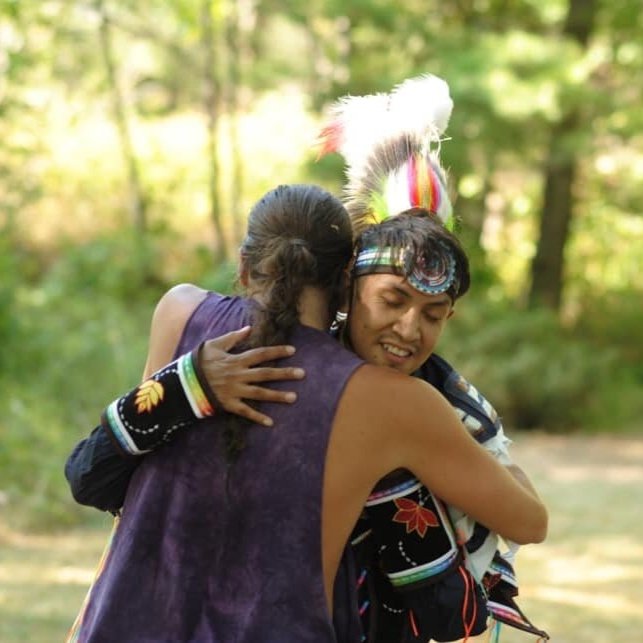 Two individuals hugging in nature. One is dressed in traditional Indigenous wear.