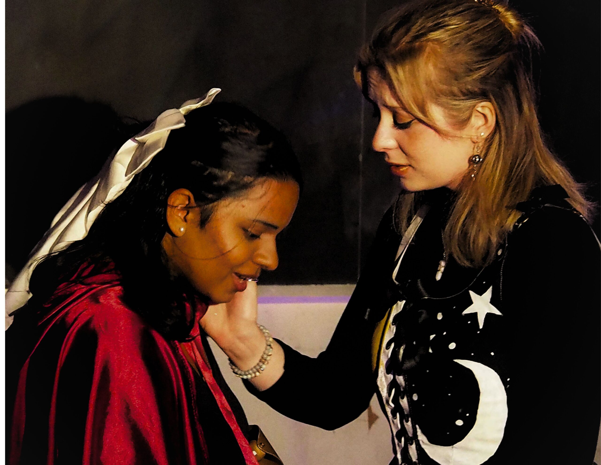 A person in a red cloak and golden hair tie speaks to a person in a black shirt with a moon and stars on it.