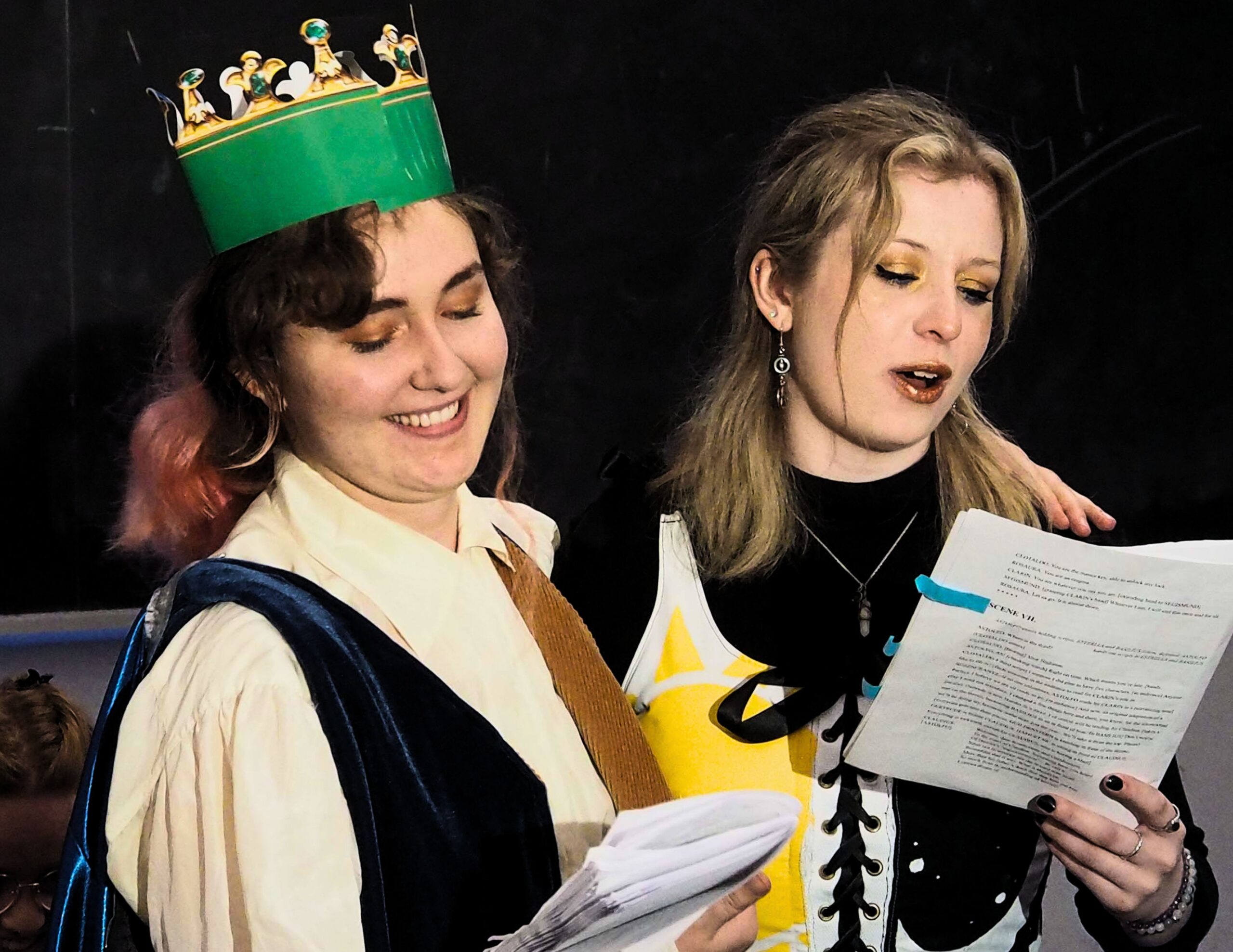 Two individuals are reading from a paper. One is wearing a paper crown. 