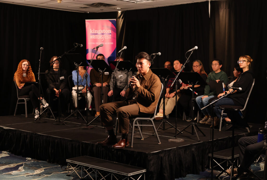 Brandon Zang and the cast of 'Ah Wing' sit on chairs on a stage with many microphones around them. 