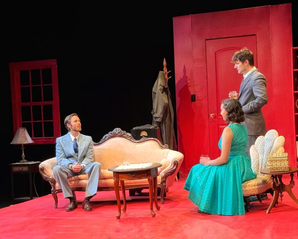 Two men and one woman appear in a red room. Each man is wearing a suit; one is standing and one is sitting on a couch. The woman sits in a chair in a turquoise dress. 
