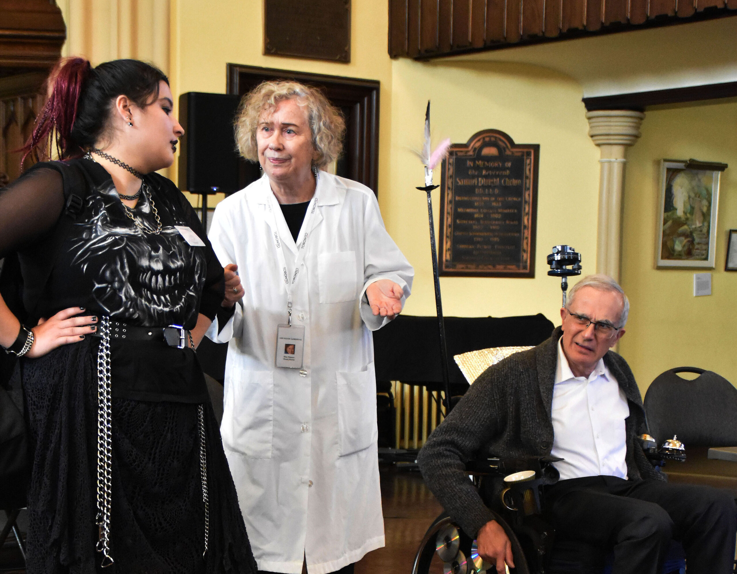 A woman dressed in black clothing, a woman dressed in a white lab coat, and a man in a wheelchair are interacting.