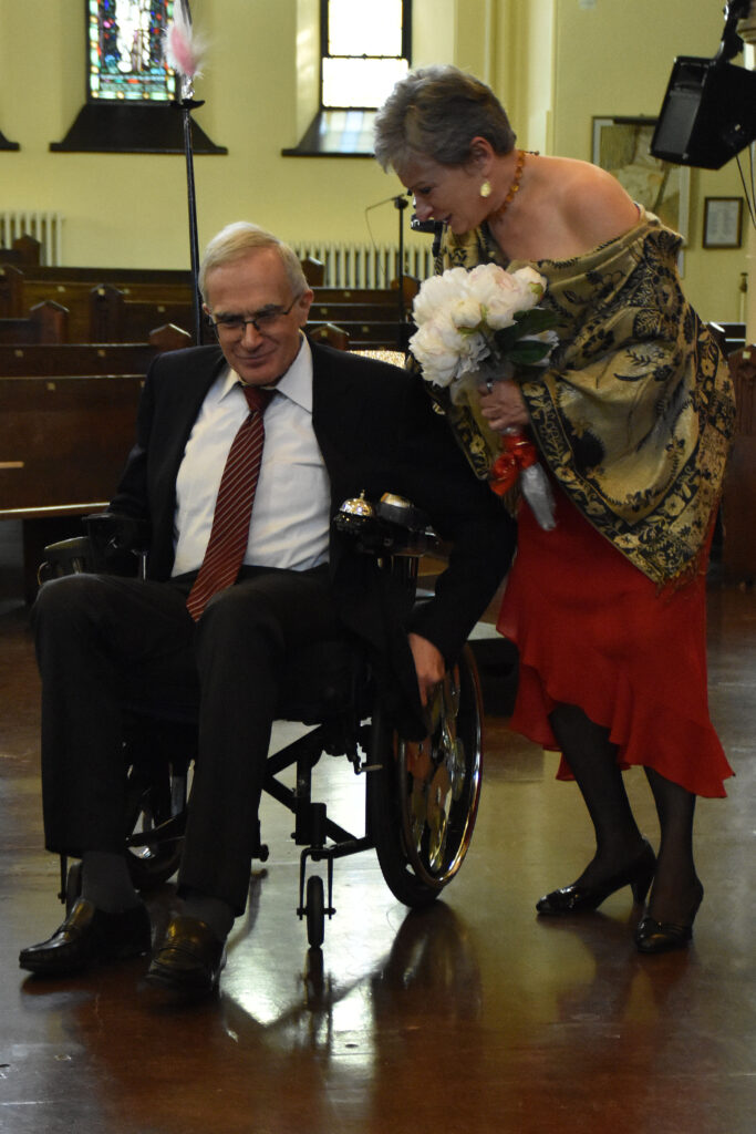 A man dressed in a suit sits in a wheelchair while a beautifully dress woman leans over and speaks to him. 