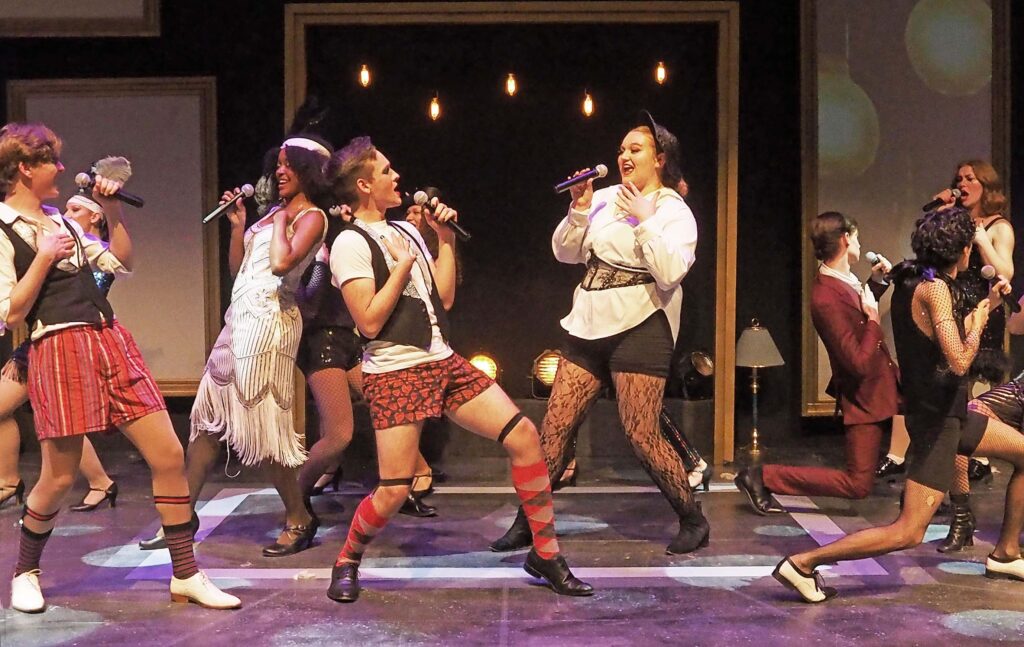 Performers on a stage singing into microphones. The costuming involves flapper dresses, corsets, and some people wearing boxer shorts. 