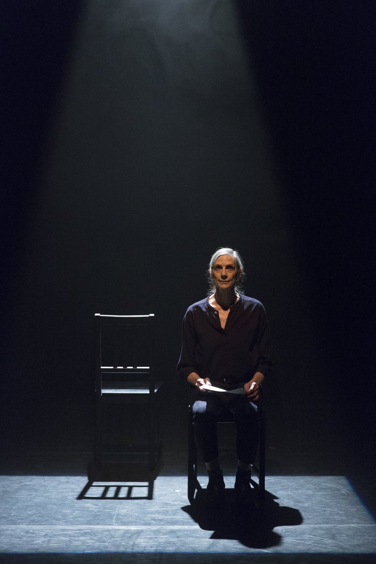 An image of a woman sitting on a stage next to an empty chair.