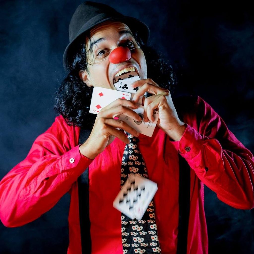 Image of a man wearing a clown nose, black hat, suspenders, and patterned tie, pulling cards from his mouth. 