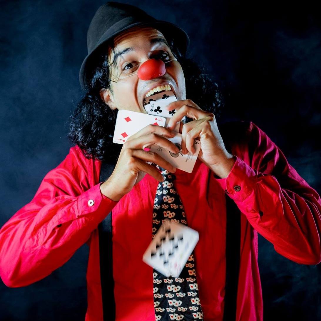 Image of a man wearing a clown nose, black hat, suspenders, and patterned tie, pulling cards from his mouth.