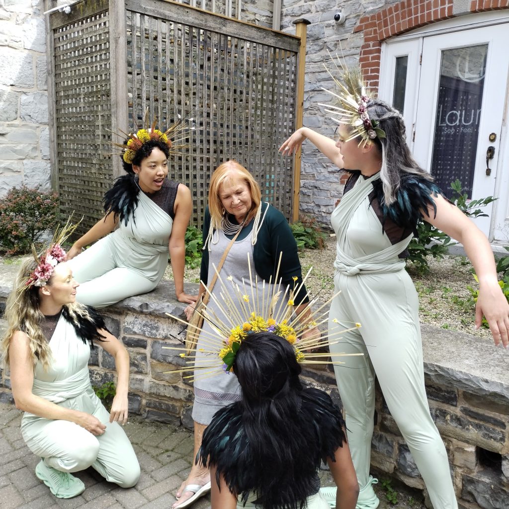 Five people outside sitting and leaning against a stone wall. Four people wear light green jumpsuits and golden headpieces. The fifth person wears a grey jumpsuit. 