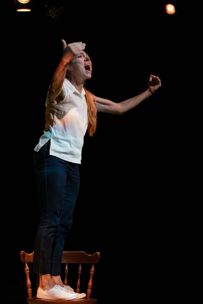 Photo of a woman standing on a chair shouting with her arms raised. 