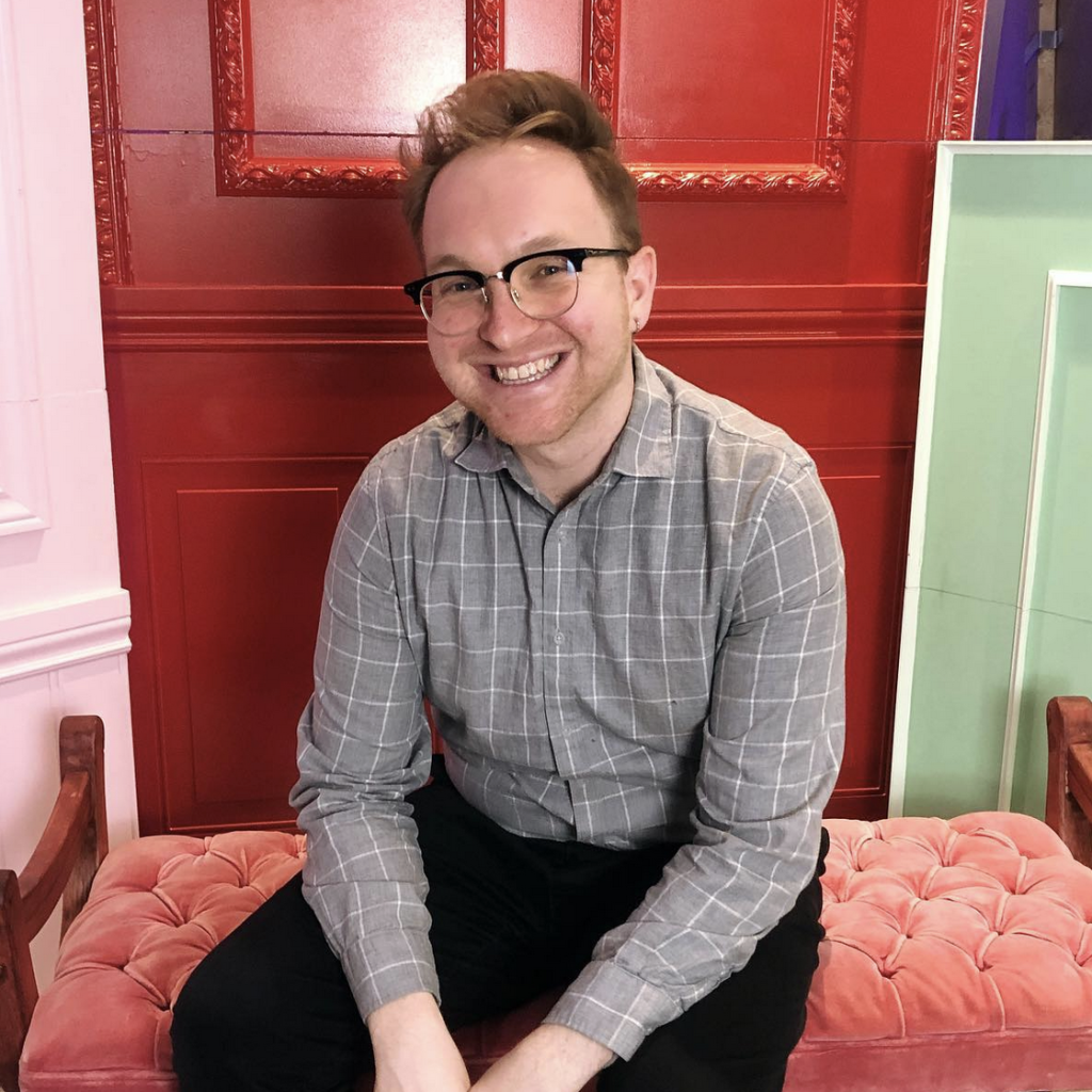 Photo of Jesse Gazic. He sits on a red bench, smiling, against a red, pink, and green wall. 