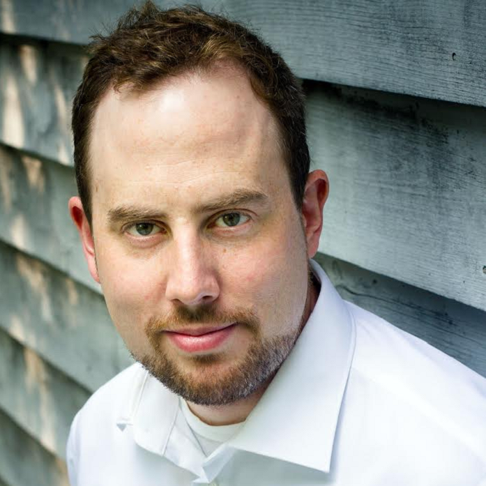 Headshot of Liam Karry. He wears a white shirt and is in front of wooden panels. 