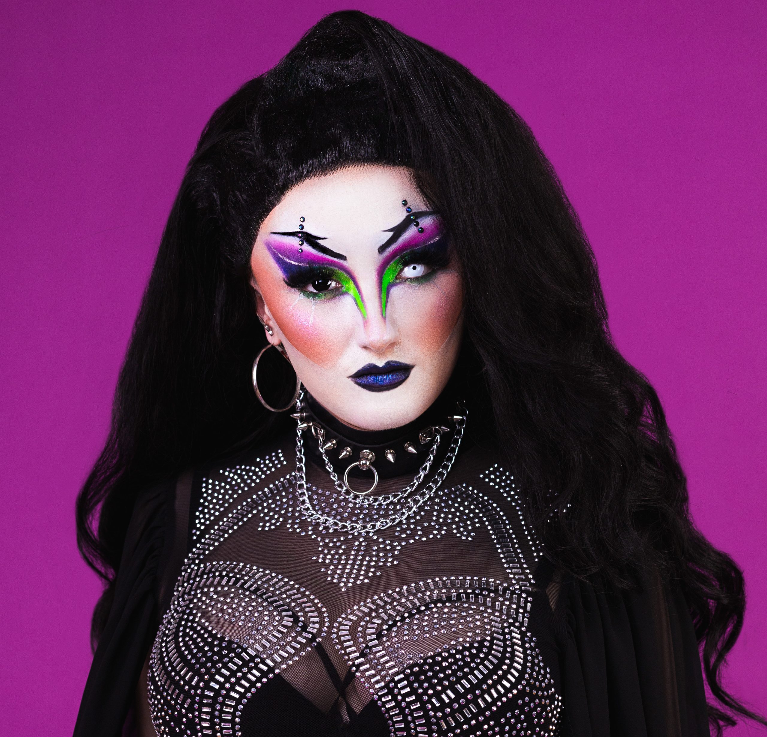 Headshot of drag queen BeeWitched. She has long black hair and wears a sparkly black bodysuit. She stands in front of a purple background.