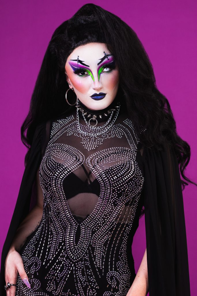 Headshot of drag queen BeeWitched. She has long black hair and wears a sparkly black bodysuit. She stands in front of a purple background. 