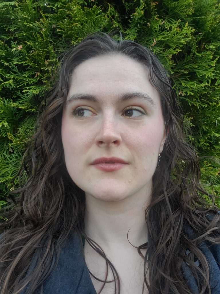 Headshot of Elsa McKnight, with long brown hair, in front of a green, leafy backdrop, looking off into the distance. 