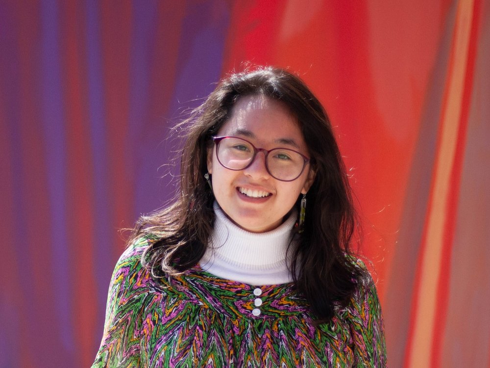 Headshot of Nicole Eun-Ju Bell. She wears a white turtleneck, with a floral shirt overtop and glasses. She is in front of a purple and red background. 