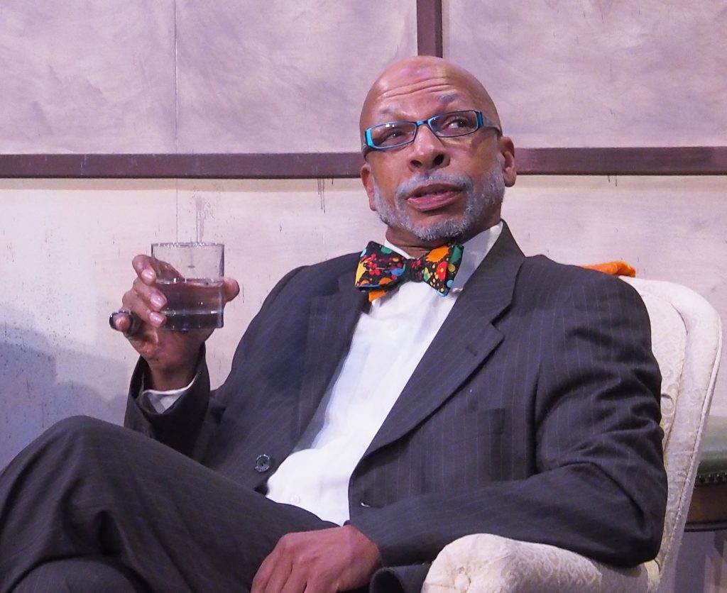 A man in a suit and colourful bowtie is sitting in a white chair. He has a drink in one hand and appears to be speaking to someone not pictured. 