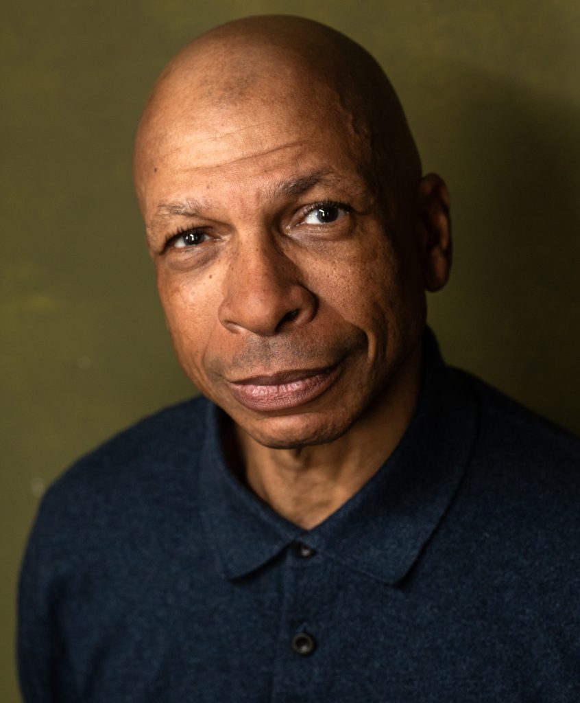 Headshot of Cassel Miles. He stares into the camera, slightly smiling. He wears a blue shirt against a dark background. 