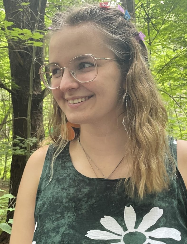 Headshot of Grace Delamere. She wears a green tank top and glasses and is smiling while looking to something off-camera. There are some trees behind here.