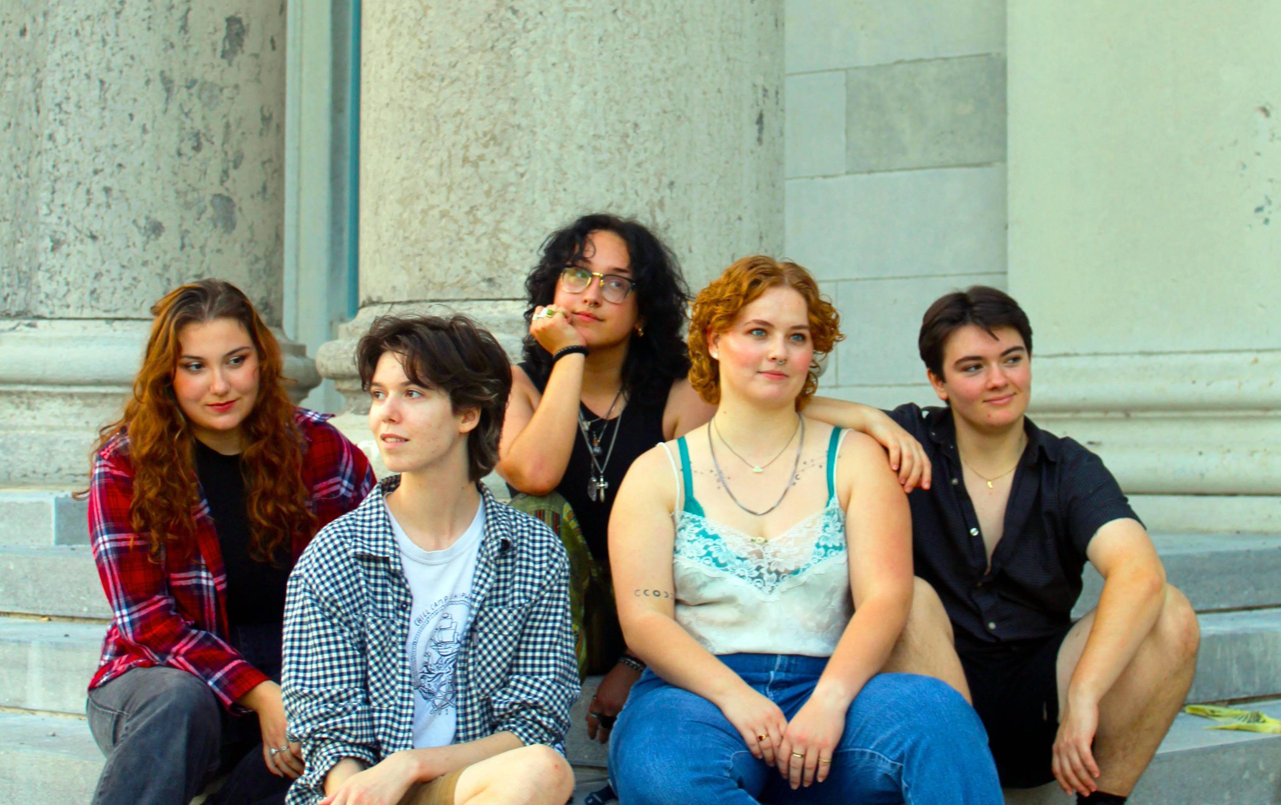 Group of five people sitting against a large white pillar. They are all dresses in casual summer clothing and looking off in different directions.