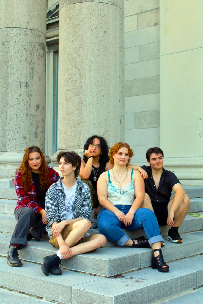 Group of five people sitting against a large white pillar. They are all dresses in casual summer clothing and looking off in different directions. 