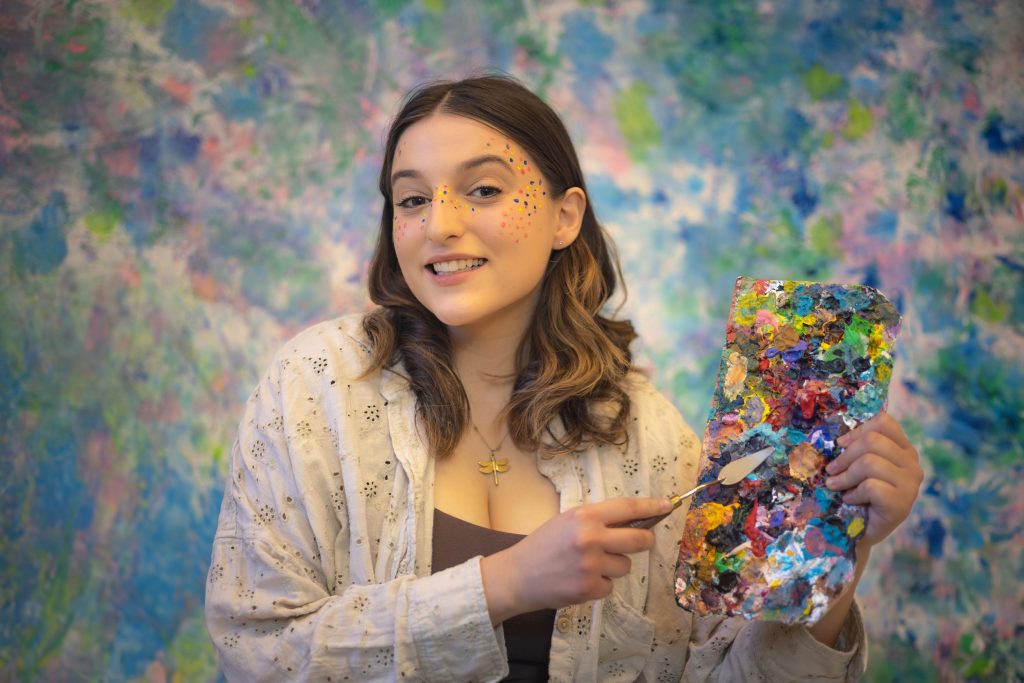 Image of a woman against a colourful painted background. She has brown hair and wears a white collared shirt over a brown shirt. She has spots of paint around her eyes and nose and smiles at the camera. She holds a paint palette covered in paint and a palette knife against the paint palette. 