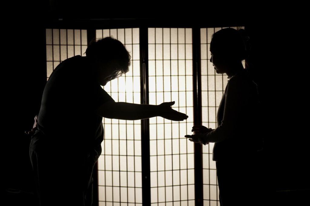 Two people stand in front of a white room divider. Light appears to come from behind the divider making the people appear as though they are shadows. One reaches out to shake the other's hand. The other has put their hand out only slightly. 