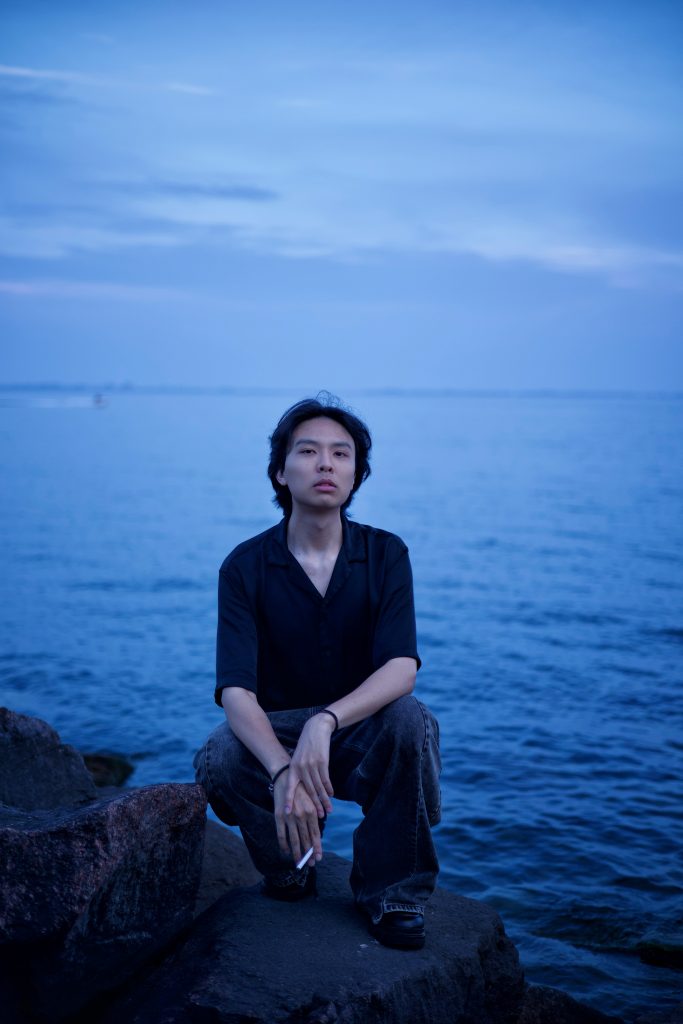 Image of a man with dark hair, wearing a dark shirt and dark pants stands low to the ground on a rock. He holds a cigarette in one hand. Deep blue water and a deep blue sky are behind him. 