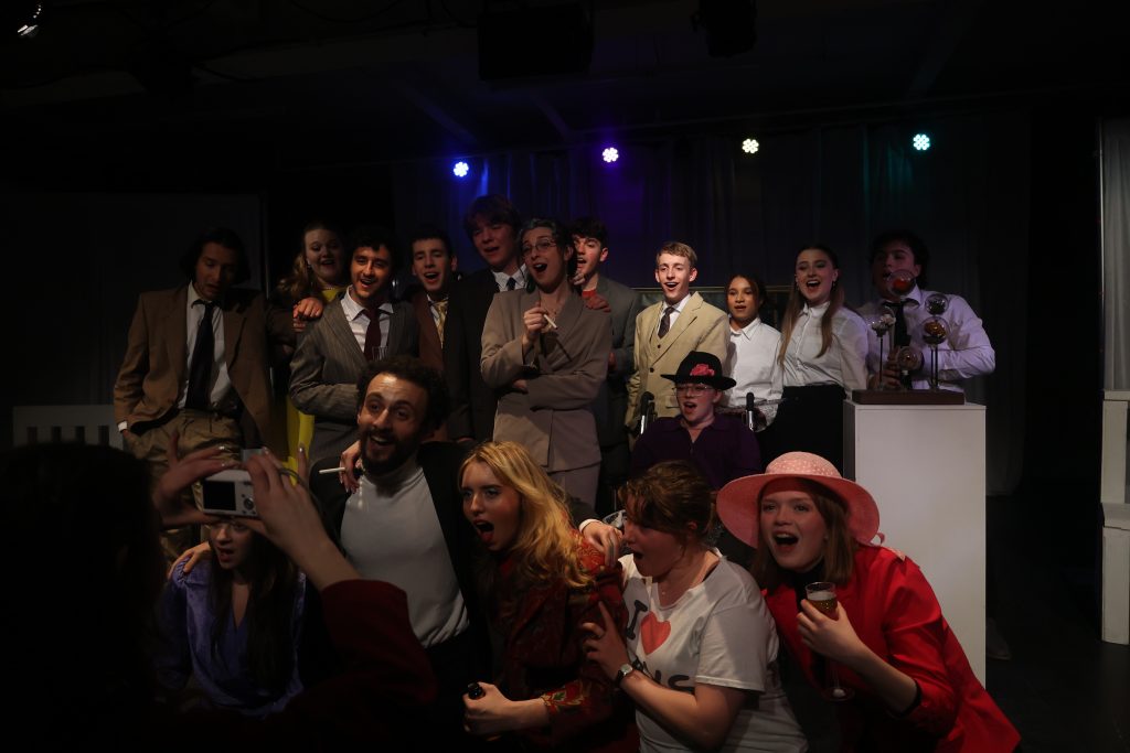 A group of performers on stage. Many are in suits, well dressed in collared or shirts while a few are in T-shirts. Some performers hold cigarettes. The cast is group together into two rows, standing or sitting right next to one another. They appear to mid-note as they face the edge of the stage and each have their mouths open ajar. 