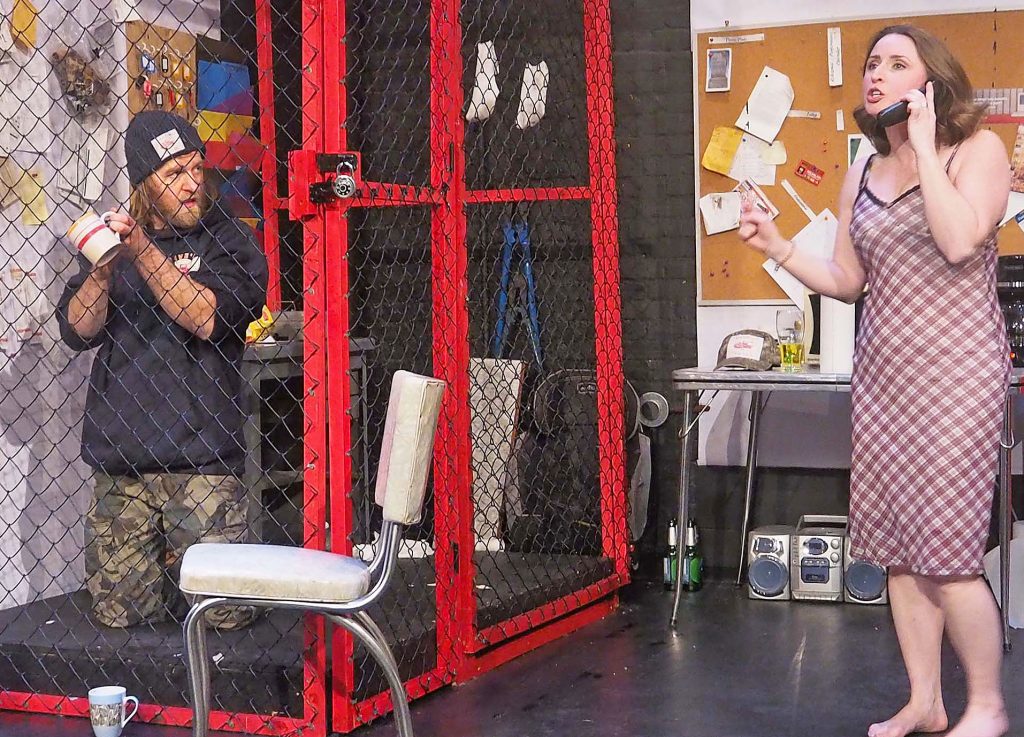 An office setting with a fenced-in portion of the office on the left. A man kneels in the caged-in portion holding a mug and looking at a woman outside the fenced-in area who is on the phone. She gestures animatedly. The office has a table, chair, keys hanging on the phone, a covered cork board and other office supplies. 