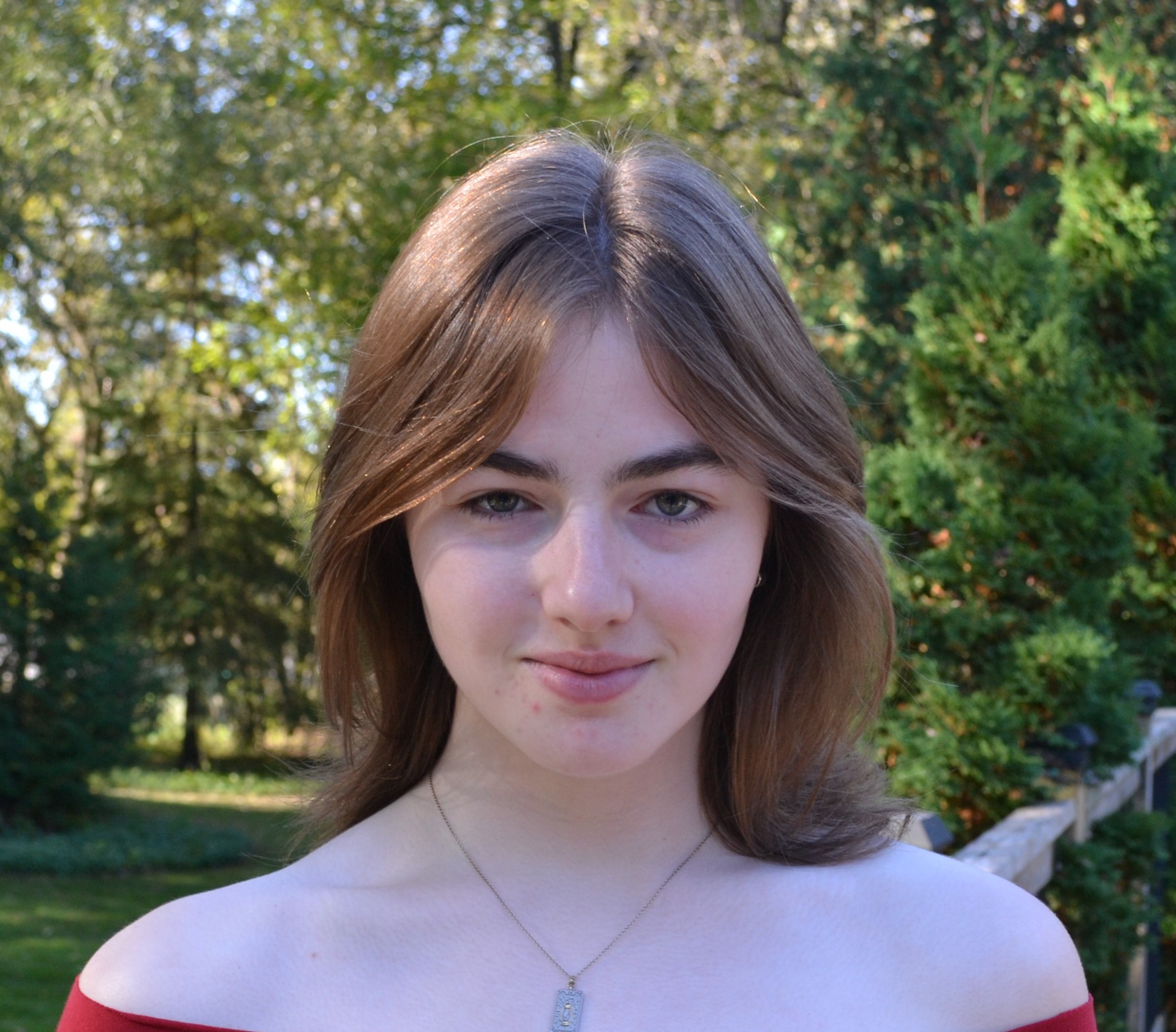 Headshot of Nic Lindegger. They wear an off-the-shoulder red top. The background is full of trees. Lindegger stares straight into the camera, slightly smiling.