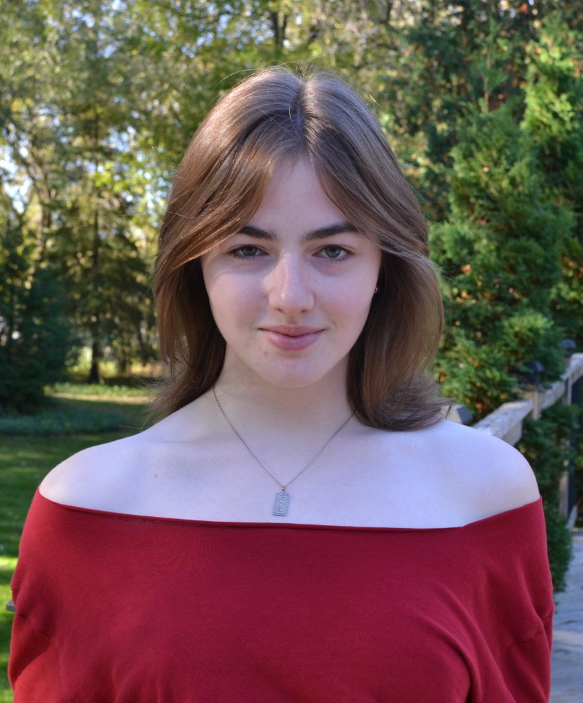 Headshot of Nic Lindegger. They wear an off-the-shoulder red top. The background is full of trees. Lindegger stares straight into the camera, slightly smiling. 