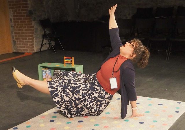 In a staged setting of a classroom, a teacher lies on a carpeted floor with one hand in the air and her feet lifted slightly off the ground.