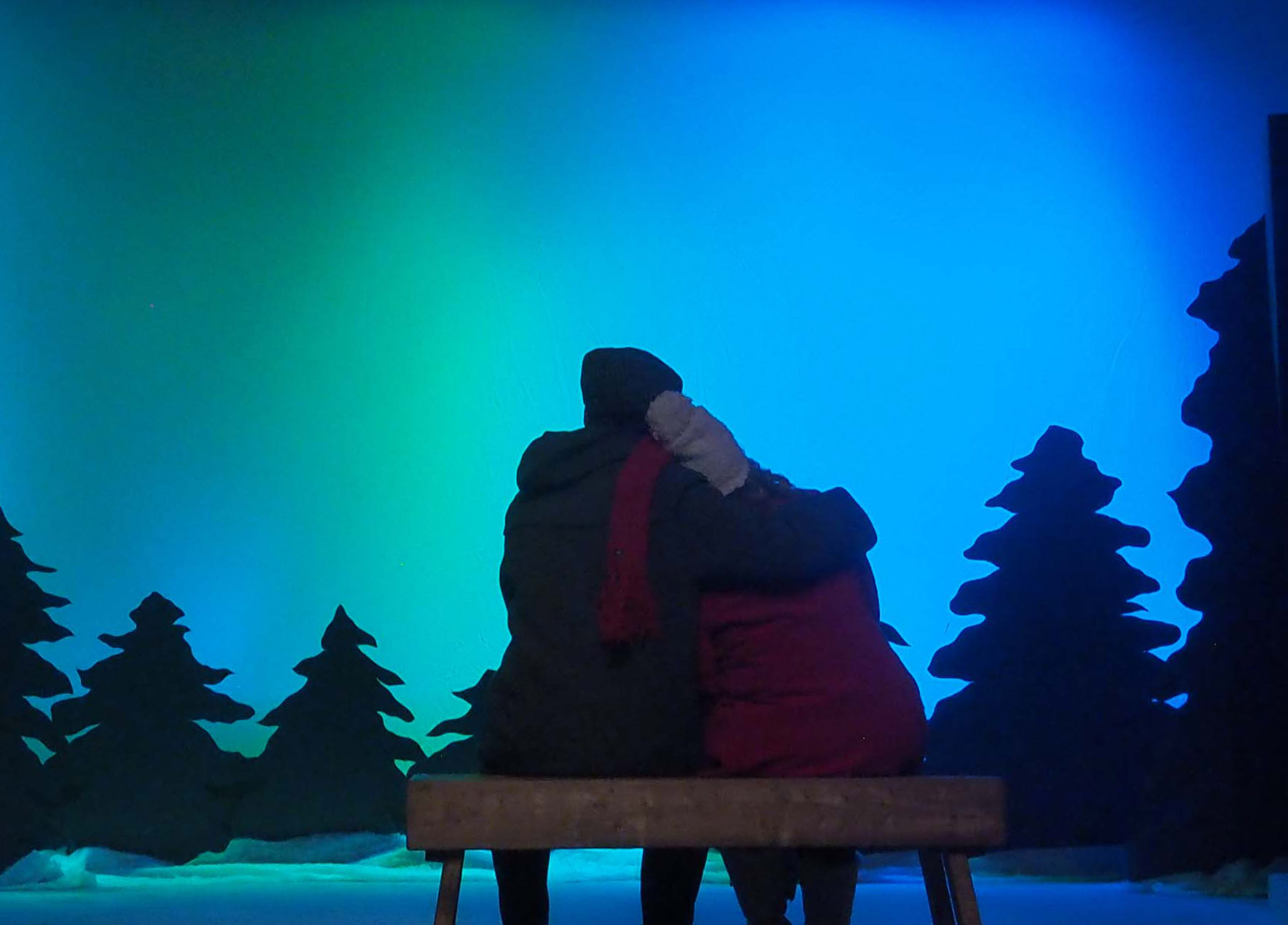 A couple sit on a bench with the backs to the camera. They're on a stage and behind them are staged trees and imagery of the northern lights.