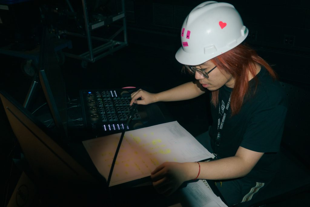 Jerall Li sits at a table with a lighting board in a dark room. He wears a white hat, glasses, and a black shirt. 