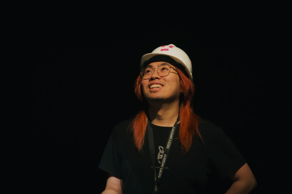 Headshot of Jerall Li looking up and smiling. He wears glasses, a white hat, a black shirt, and is against a black background. 