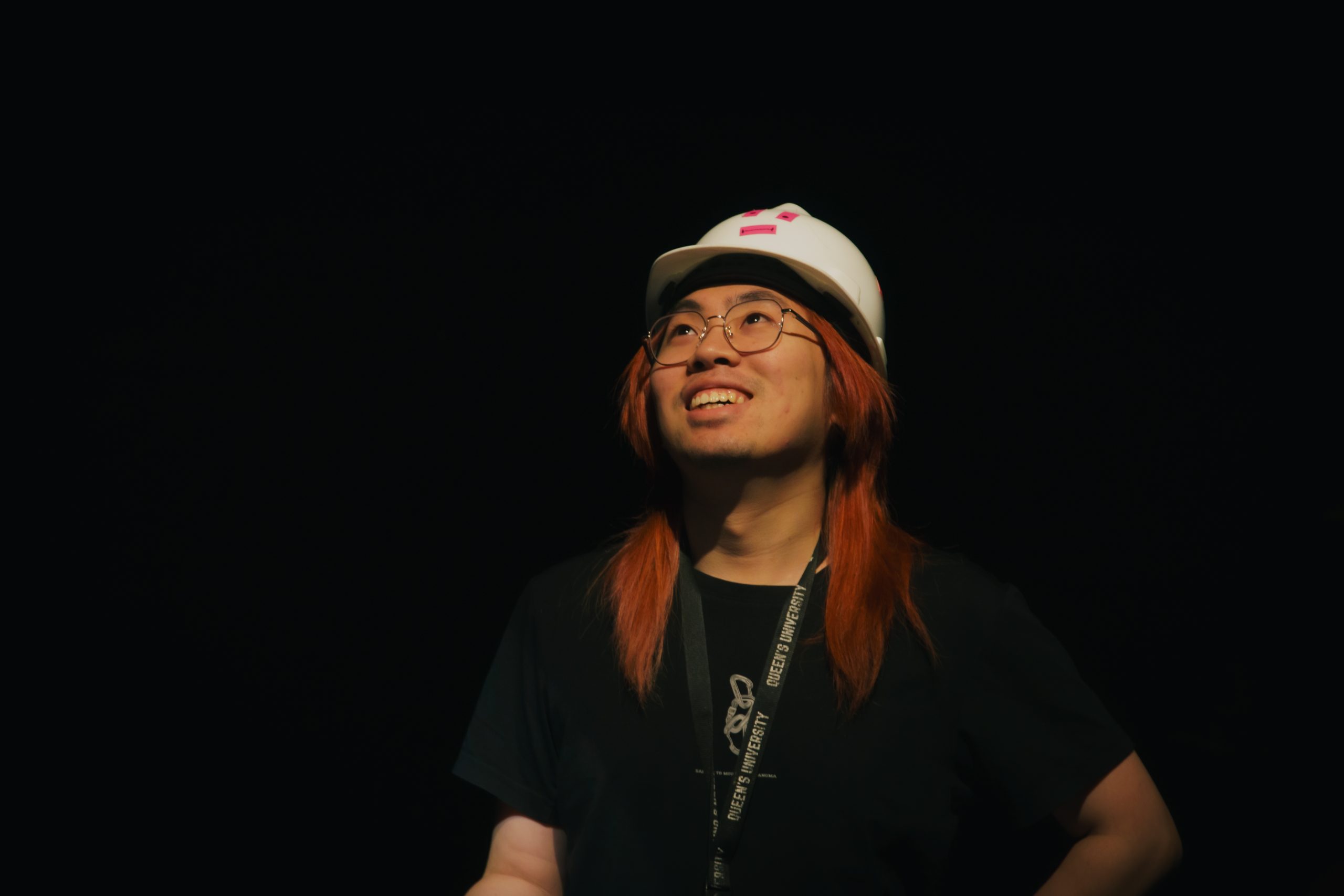 Headshot of Jerall Li looking up and smiling. He wears glasses, a white hat, a black shirt, and is against a black background.