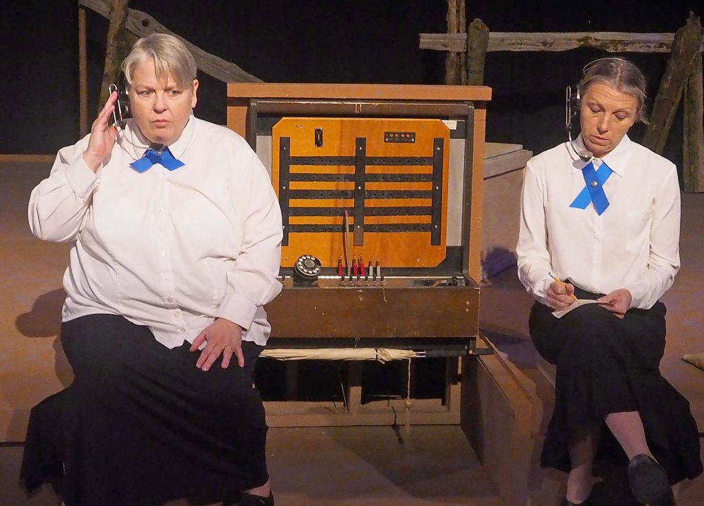 Two actors wear headsets and sit on either side of a wooden box. One touches their headset and the other is writing something donw.