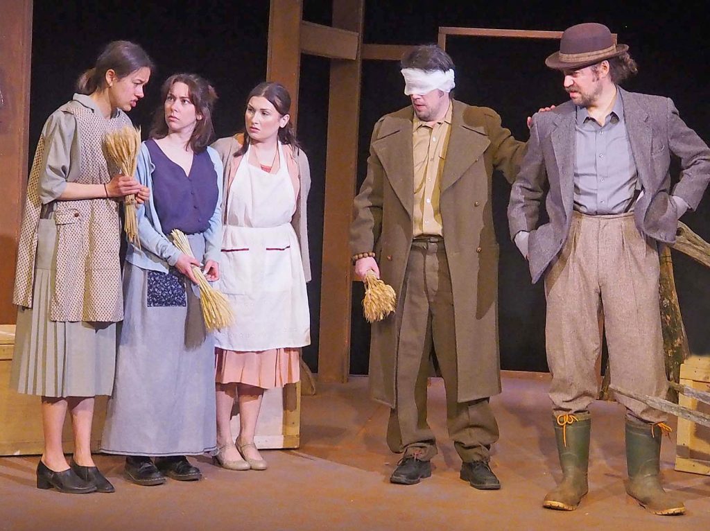 Five actors are on stage and speaking with each other. A few hold bundles of hay. One has gauze over their eyes.
