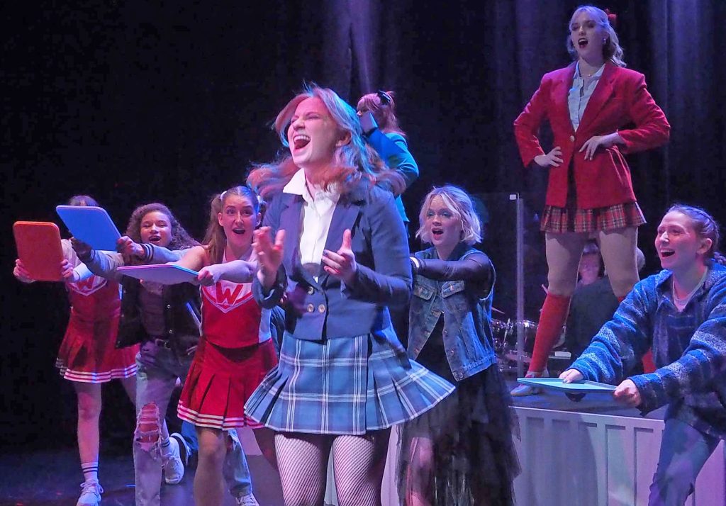 Performers on stage. Some wear school uniforms and some wearing cheerleading uniforms. The front and centre performer is overjoyed and jumps gleefully.